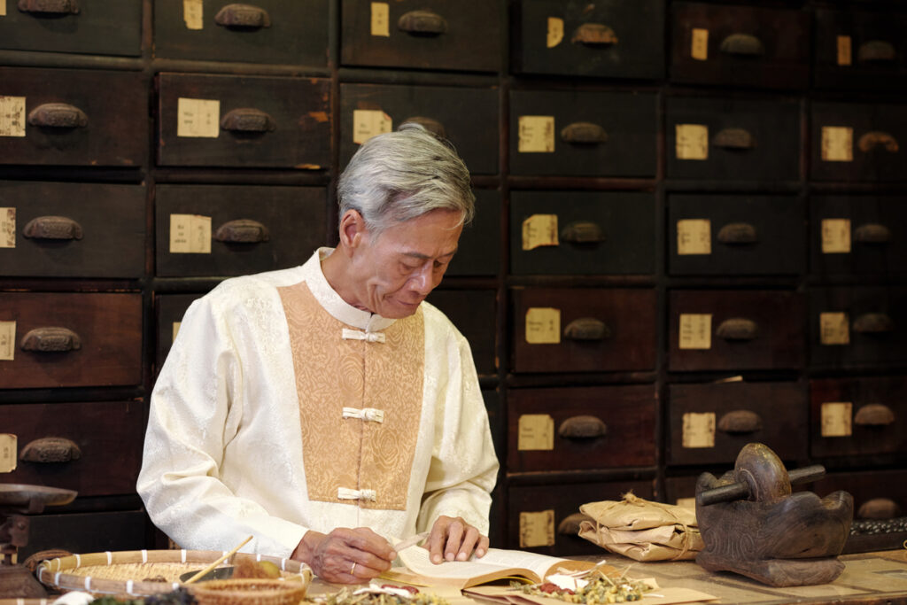 Asian Apothecary Examining Herbs In Handbook - tstpp Аптекарь изучает травы в справочнике