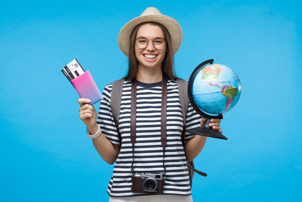 Жизнерадостная девушка держит в одной руке глобус, а в другой — паспорт с авиабилетами
