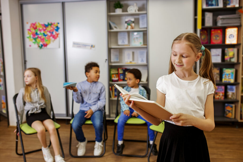 Школьница стоит посреди класса и читает что-то из книги