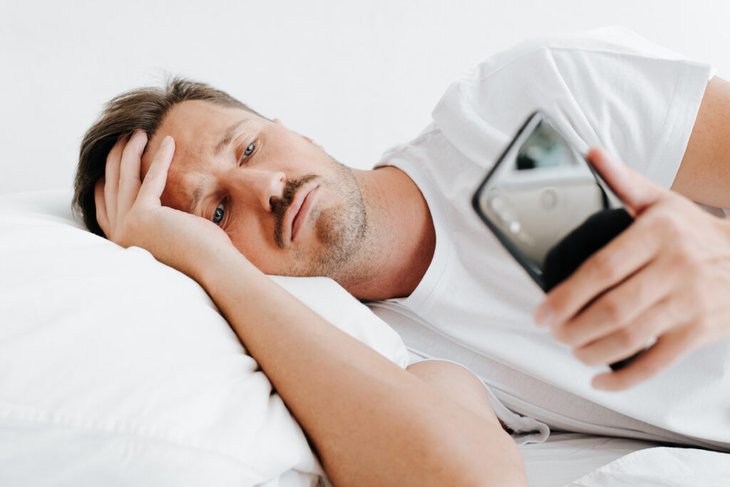 Мужчина лежит в постели и устало смотрит в телефон
