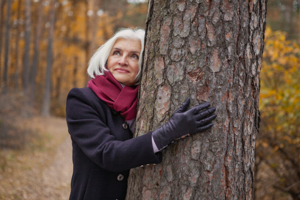 Пожилая женщина в красивом пальто стоит у дерева в осеннем лесу