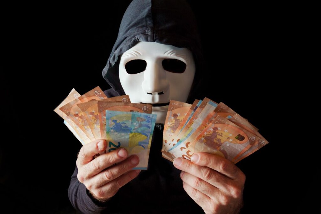 хакер с белой маской и черным капюшоном, считающий банкноты