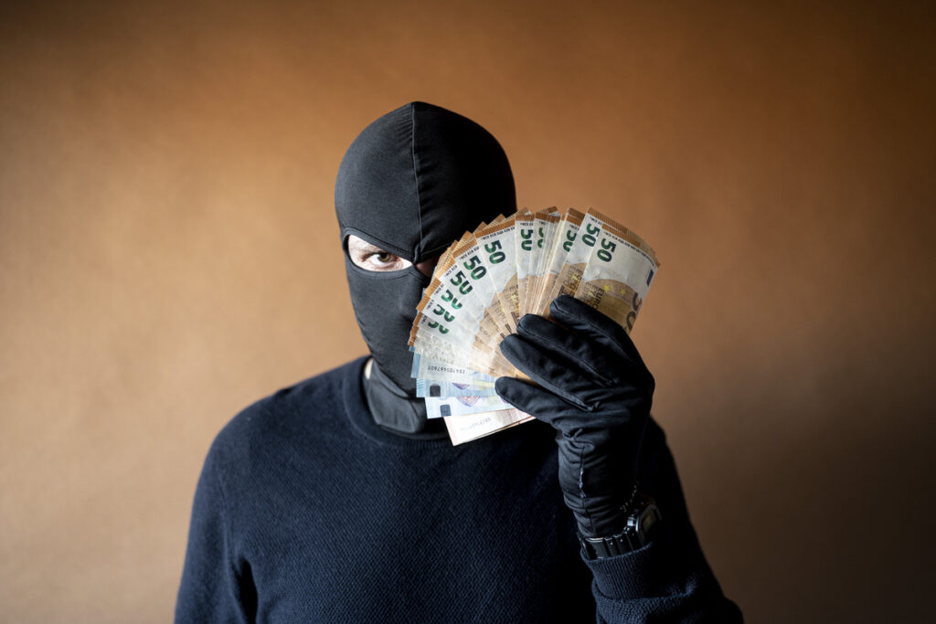 мужчина-вор с балаклавой на голове, держащий перед глазами веер из купюр