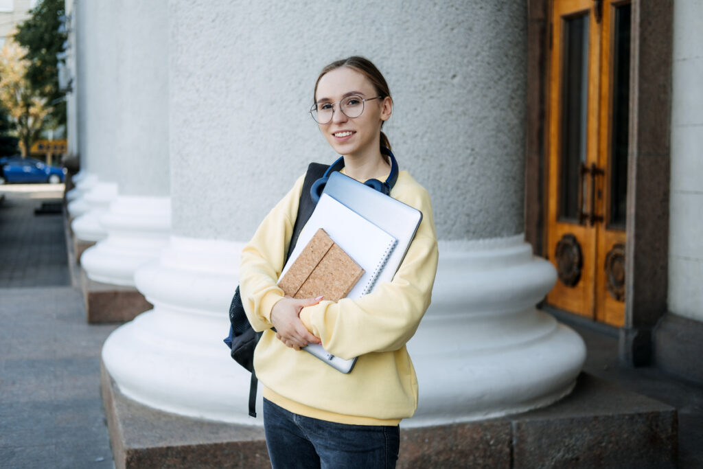Счастливая студентка колледжа с ноутбуком и книгами возле колледжа