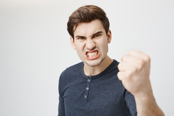 Fierce confident stylish european dark-haired male with trendy haircut holding fist in front of him as if ready for fight or challenge, clenching white teeth, having aggressive expression on face Мужчина держит перед собой кулак