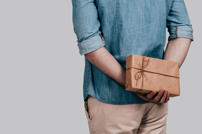 Surprising her... Close-up rear view of man in blue jeans shirt holding a gift box behind his back while standing against grey background Мужчина держит подарок за спиной