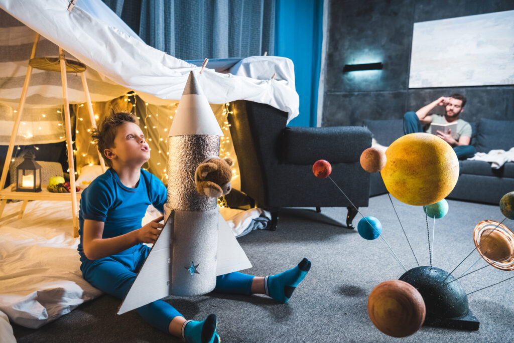 boy sitting in blanket fort with toy rocket and solar system model father sitting on sofa behind - mynewton ребенок играет с ракетой в своей космической комнате