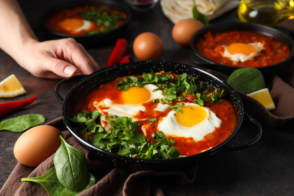 Сковороды с шакшукой, яйцами, базиликом, миской перца и масла, мужская рука на темном фоне