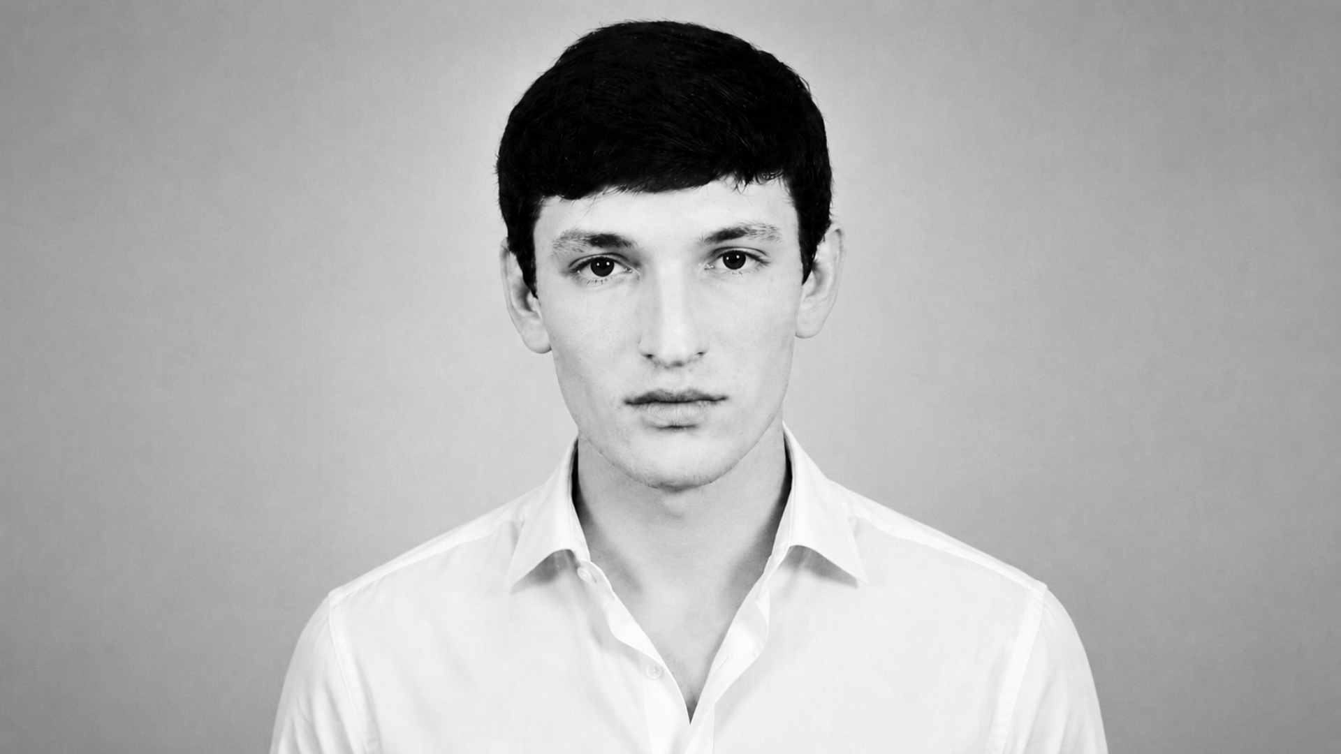 Black and white cinematic studio portrait of a man in a white shirt, no tie, with the top button ...