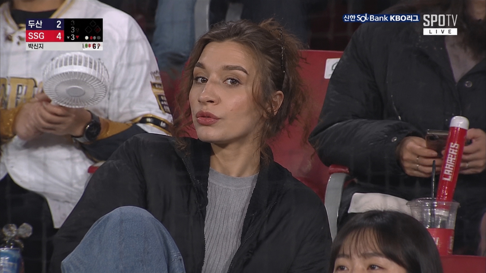 Realistic sports broadcast screenshot-style documentary photo set in the spectator stands of a Ko...
